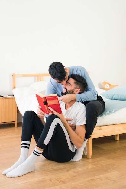 Hombre sentado en la cama besando a su novio sosteniendo un libro en la mano