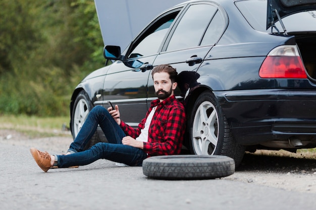 Foto gratuita hombre sentado en la carretera y apoyándose en el coche