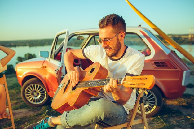 Hombre sentado y descansando en la playa tocando la guitarra en un día de verano cerca del río. vacaciones, viajes, concepto de verano.