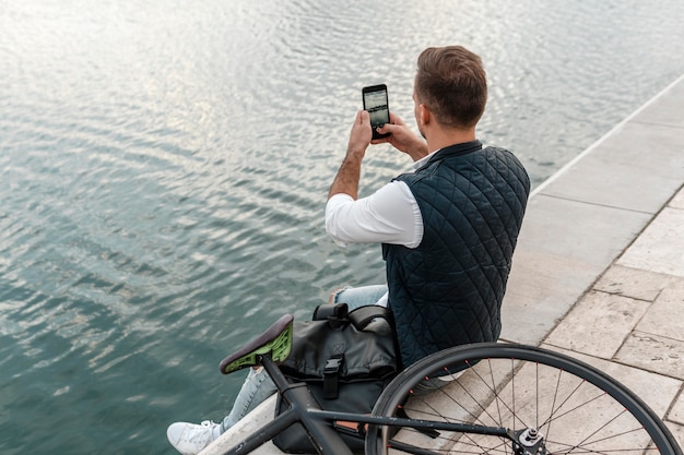 Hombre sentado junto a su bicicleta y tomando una foto de un lago