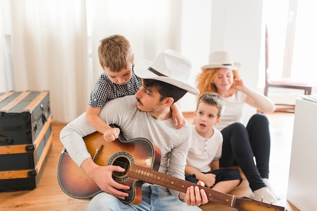 Hombre sentado en el piso de madera con su familia tocando la guitarra