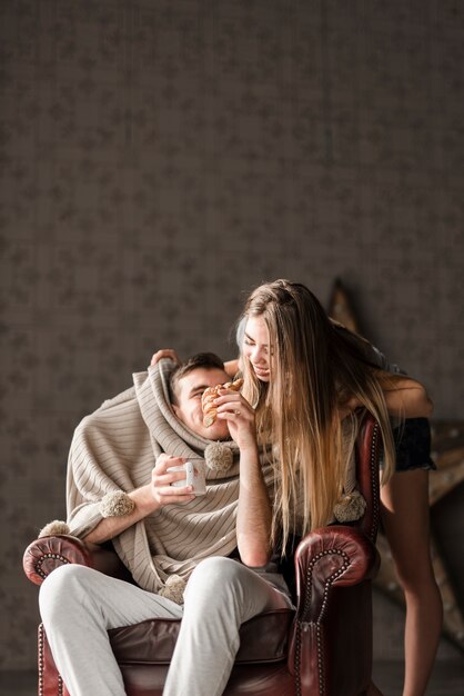 Hombre sentado en una silla que alimenta un croissant a su novia de pie detrás de él