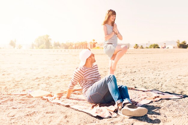 Hombre sentado sobre una manta mirando a su novia haciendo yoga en la playa