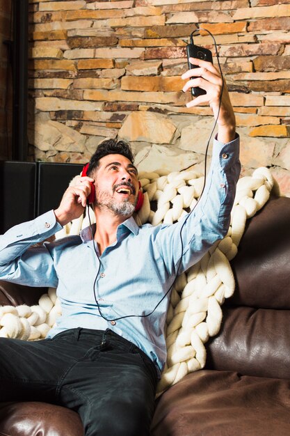 Hombre sentado en el sofá con auriculares en sus oídos haciendo una videollamada por teléfono inteligente