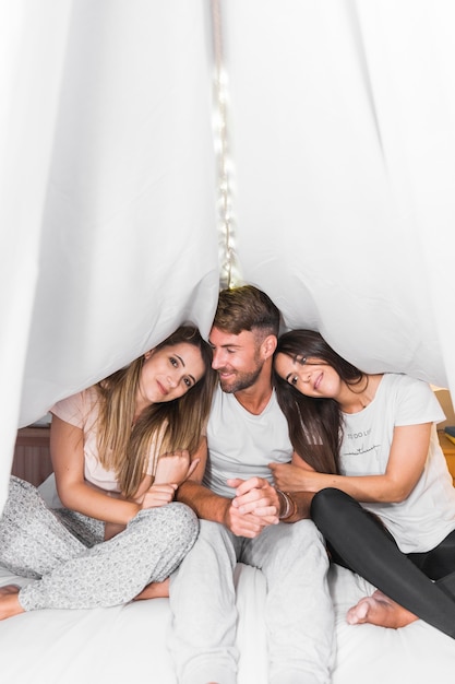 Hombre sentado con sus dos amigas en la cama bajo la cortina blanca