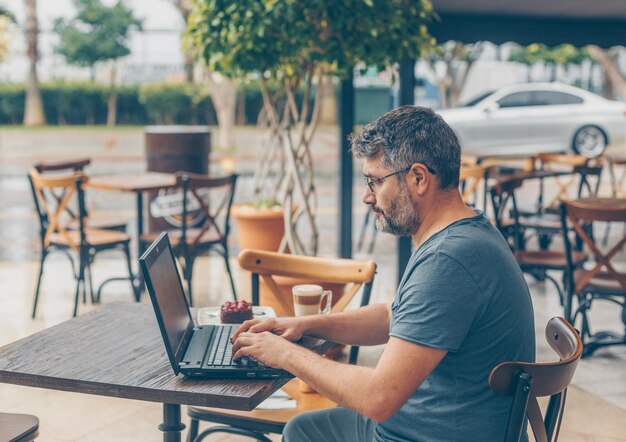 Hombre sentado y trabajando en la computadora portátil en la terraza del café durante el día y mirando ocupado