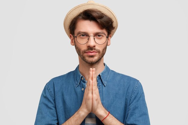 Hombre serio sin afeitar con expresión facial tranquila, se para en gesto de oración, pide perdón, vestido con camisa de mezclilla