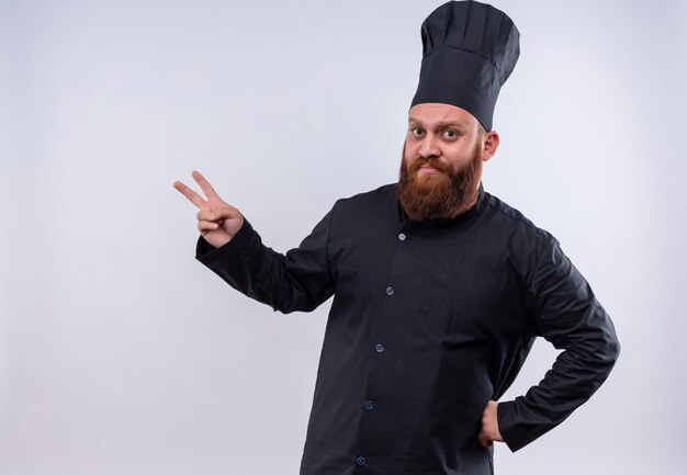 Un hombre serio chef barbudo en uniforme negro que muestra el gesto de las dos manos mientras mira en una pared blanca
