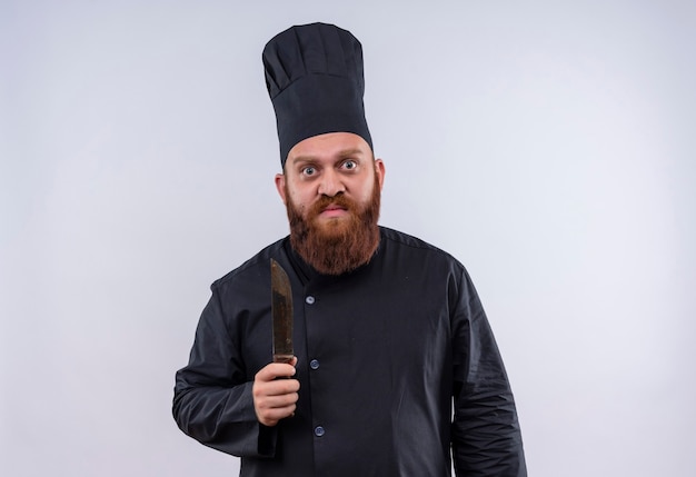 Un hombre serio chef barbudo en uniforme negro sosteniendo un cuchillo mientras mira a la cámara en una pared blanca