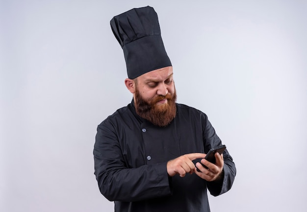 Un hombre serio chef barbudo en uniforme negro tocando su teléfono móvil en una pared blanca