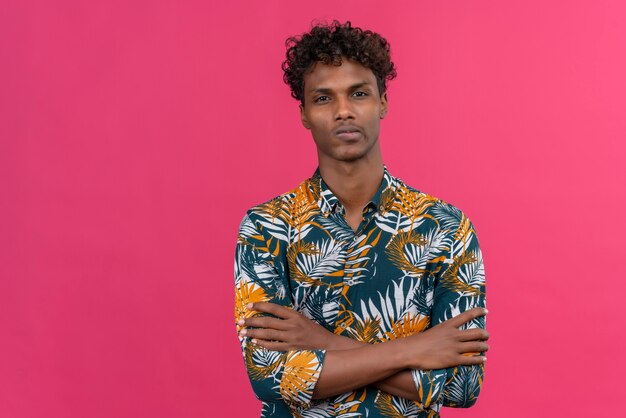 Hombre serio confiado de piel oscura con cabello rizado en hojas camisa estampada tomados de la mano doblados mientras mira a cámara sobre un fondo rosa