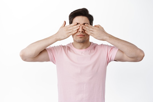 El hombre serio cubre los ojos con las manos y espera algo, anticipando la sorpresa, de pie contra la pared blanca