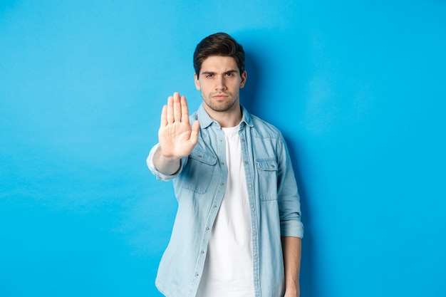 Hombre serio frunciendo el ceño y diciendo que no, extendiendo la mano a la señal de stop de la tienda, prohíbe la acción, de pie contra el fondo azul