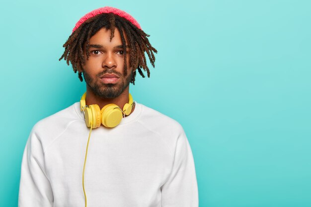 Hombre serio hipster con rastas, viste sombrero rosa y suéter blanco, tiene auriculares para escuchar pistas de audio, pasa tiempo libre siempre escuchando música