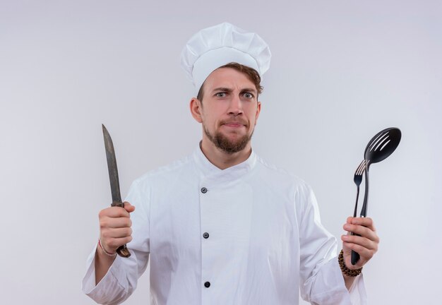 Un hombre serio joven chef barbudo vestido con uniforme de cocina blanco y sombrero sosteniendo cuchillo, tenedor y cucharón mientras mira en una pared blanca