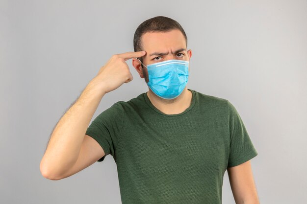 Hombre serio joven que lleva la máscara médica de la cara que señala a su cabeza con el dedo aislado en blanco