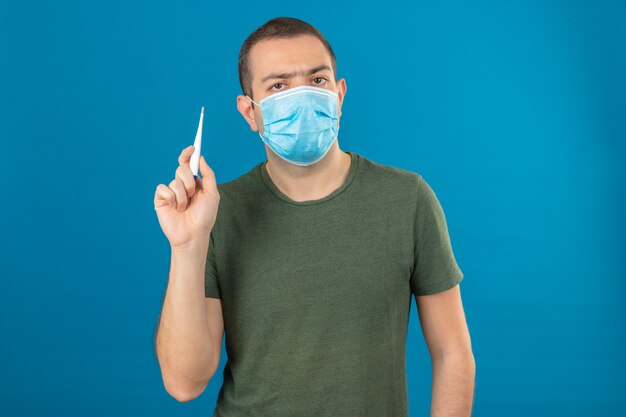 Hombre serio joven que lleva la máscara médica de la cara que sostiene un termómetro digital en levantar la mano aislada en azul