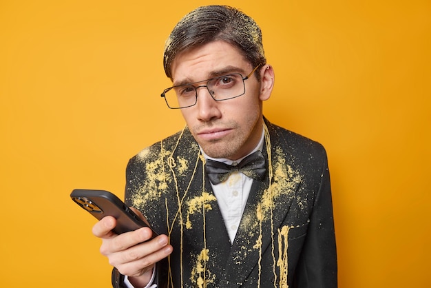 El hombre serio mira con sospecha usa el teléfono móvil para chatear en línea vestido con ropa festiva celebra una ocasión especial pasa tiempo en una fiesta de swag aislada sobre un fondo amarillo vívido
