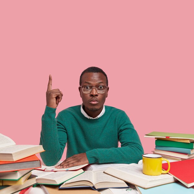 Hombre serio nerd usa grandes gafas, suéter verde, apunta hacia arriba con un dedo, rodeado de muchos libros mientras se prepara para la sesión de examen