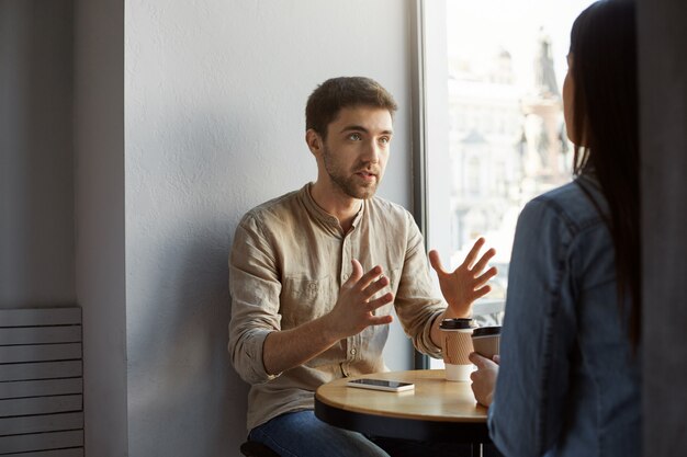 Hombre serio de pelo oscuro sin afeitar sentado en la cafetería con el cliente, hablando y gesticulando con las manos, tratando de aclarar algunos detalles de la comisión que recibió.