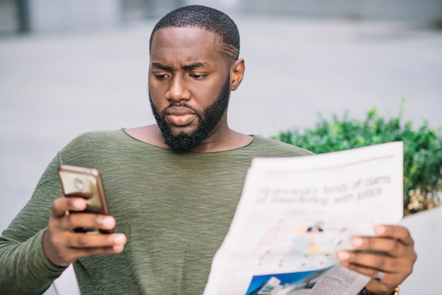 Foto gratuita hombre serio con periódico usando teléfono inteligente