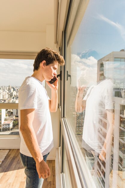 Hombre serio de pie delante de la ventana de cristal mientras habla por teléfono móvil