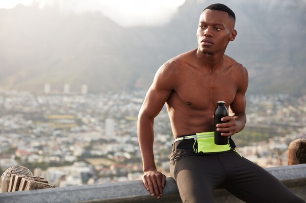 El hombre serio de piel oscura practica deportes extremos en las montañas, sostiene una botella con una bebida fresca, está absorto en sus pensamientos, piensa en metas futuras, lleva un estilo de vida activo y saludable. Modelo masculino de fitness.