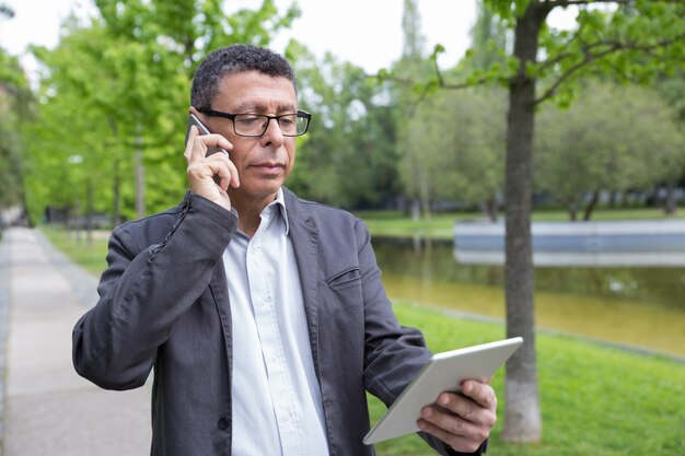 Hombre serio que hojea en la tableta y que habla en el teléfono en parque