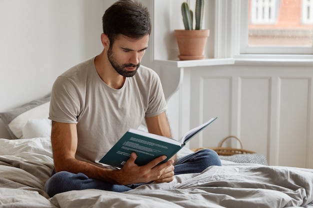 Hombre serio y relajado sin afeitar sostiene el libro delante de la cara, vestido con una camiseta informal, se sienta en la postura del loto en la cama