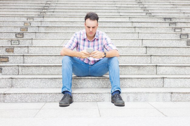 Hombre serio sentado en las escaleras y mensajes de texto en el teléfono