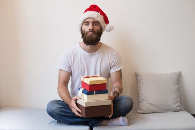 Hombre serio con sombrero de Santa y sosteniendo cajas de regalo en el sofá