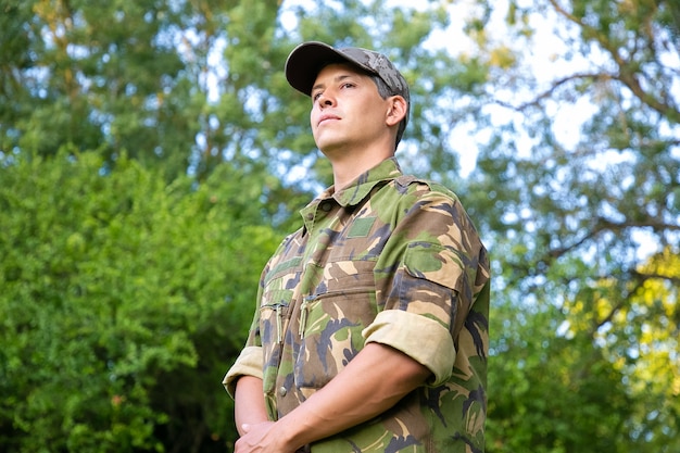 Hombre serio en uniforme de camuflaje militar de pie en el parque, mirando a otro lado.