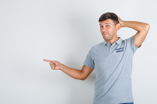 Foto gratuita hombre de servicio técnico apuntando al lado con la mano en la cabeza en camiseta gris y mirando tímido