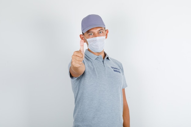 Hombre de servicio técnico mostrando el pulgar hacia arriba en camiseta gris con gorra y máscara médica y mirando con cuidado