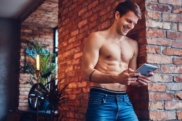 Un hombre sexy sin camisa tatuado con touchpad se inclina contra la pared de ladrillo. Sonriendo y mirando a otro lado.