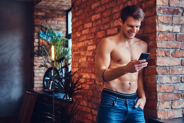 Un hombre sexy sin camisa tatuado usando un teléfono móvil se inclina contra la pared de ladrillo. Sonriendo y mirando a otro lado.