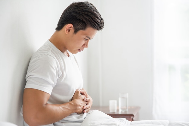 Un hombre se sienta en la cama con dolor de estómago y se presiona el estómago con las manos.