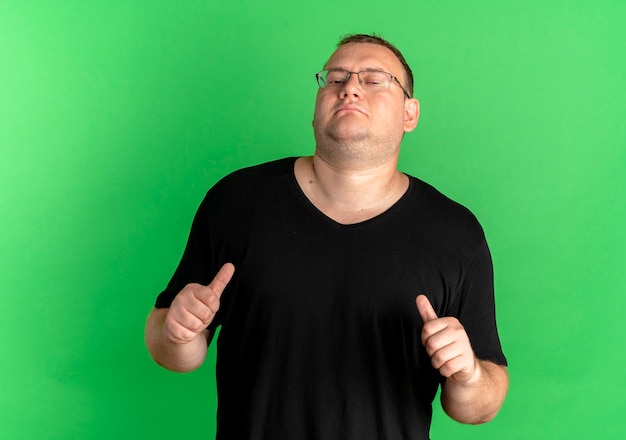 Foto gratuita hombre con sobrepeso complacido en gafas con camiseta negra apuntando a sí mismo de pie sobre la pared verde
