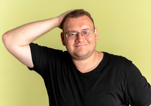 Hombre con sobrepeso complacido en gafas con camiseta negra mirando a la cámara sonriendo de pie sobre la pared de luz