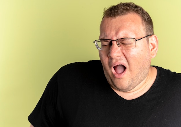 Foto gratuita hombre con sobrepeso decepcionado con gafas con camiseta negra con la boca abierta de pie sobre la pared de luz