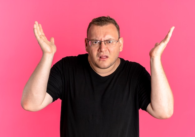 Hombre con sobrepeso en gafas con camiseta negra confundido y disgustado con los brazos levantados como preguntando o discutiendo de pie sobre una pared rosa