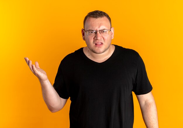 Hombre con sobrepeso en gafas con camiseta negra disgustado con el brazo extendido como preguntando o discutiendo sobre naranja