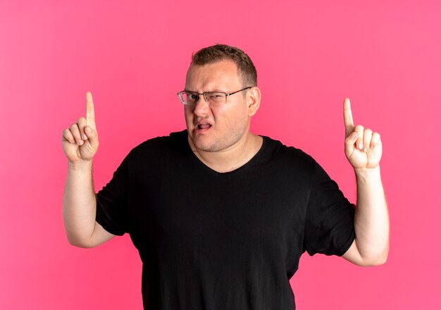 Hombre con sobrepeso en gafas con camiseta negra disgustado mostrando los dedos índices de pie sobre la pared rosa