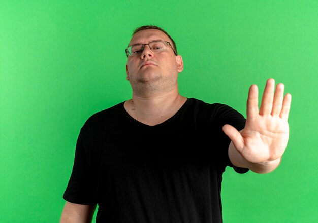 Foto gratuita hombre con sobrepeso en gafas con camiseta negra haciendo dejar de cantar con la mano abierta con cara seria de pie sobre la pared verde