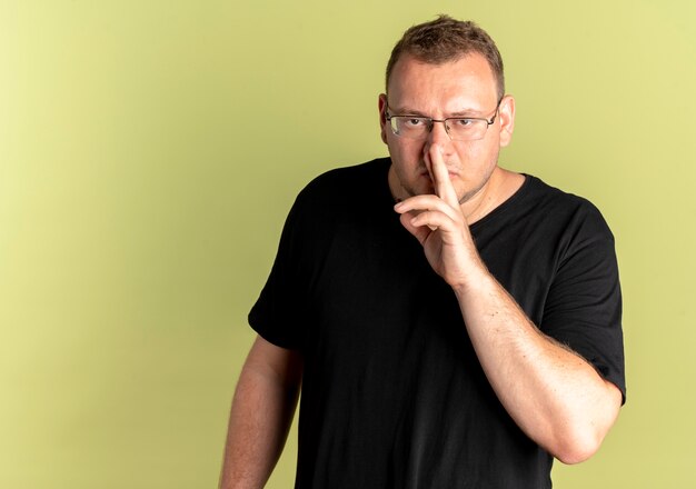 Hombre con sobrepeso en gafas con camiseta negra haciendo gesto de silencio con el dedo en los labios de pie sobre la pared de luz