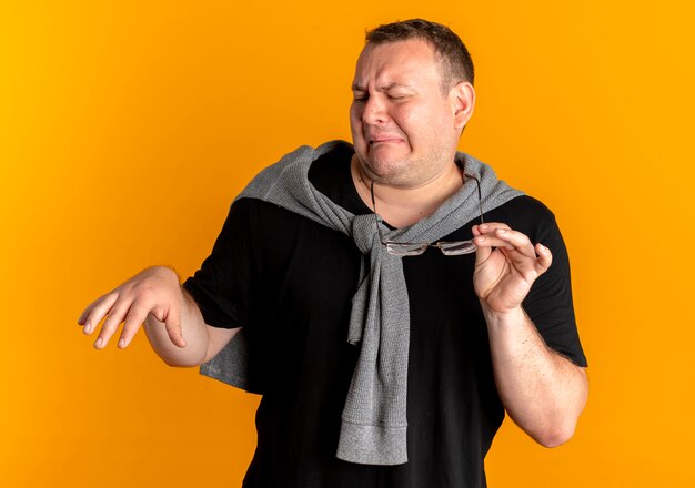 Hombre con sobrepeso en gafas con camiseta negra haciendo gestos de defensa con las manos con expresión de disgusto de pie sobre la pared naranja