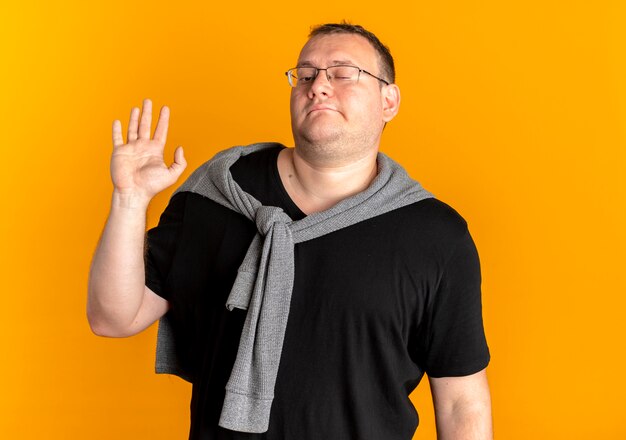 Hombre con sobrepeso en gafas con camiseta negra mirando confiado saludando con la mano de pie sobre la pared naranja