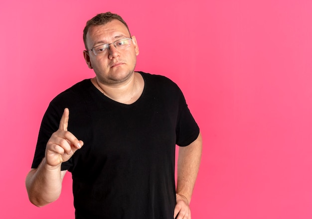 Hombre con sobrepeso en gafas con camiseta negra mirando a un lado con expresión triste en la cara mostrando el dedo índice de pie sobre la pared rosa