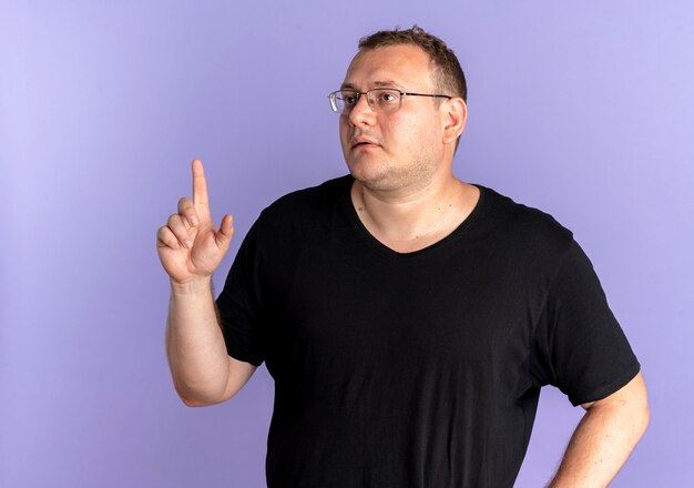 Hombre con sobrepeso en gafas con camiseta negra mirando a un lado mostrando el dedo índice pidiendo esperar un minuto sobre azul