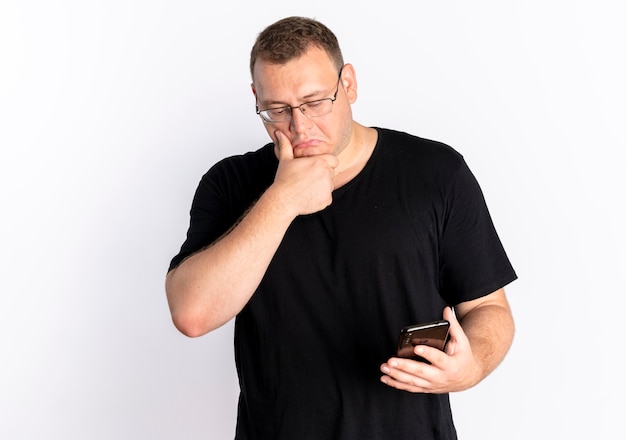 Hombre con sobrepeso en gafas con camiseta negra mirando la pantalla de su teléfono inteligente desconcertado de pie sobre la pared blanca